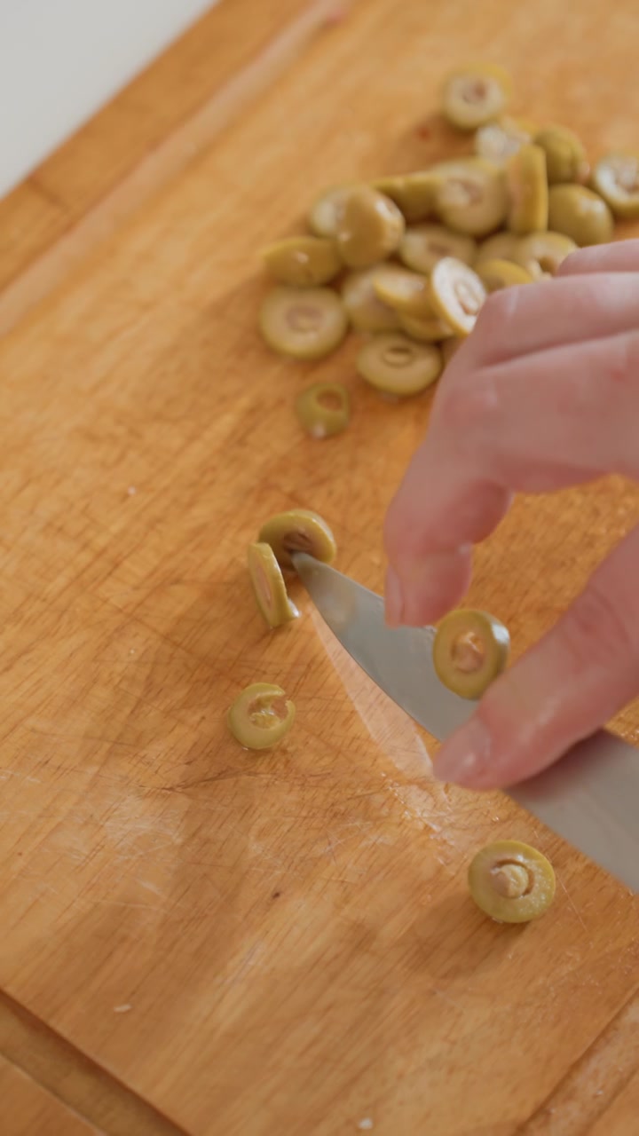 Primo piano dall'alto mostra una mano che affetta una piccola erba verde in rondelle sottili su un tagliere di legno, poi raccoglie i pezzi in un mucchietto ordinato accanto a un coltello appoggiato durante un'attenta preparazione in cucina