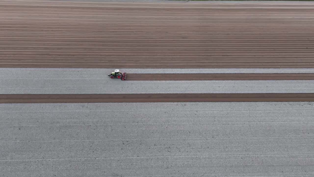 In golden-hour light slow-motion drone pan wide over tractor pulling harrow to smooth pale and dark soil furrows across vast field, at golden hour