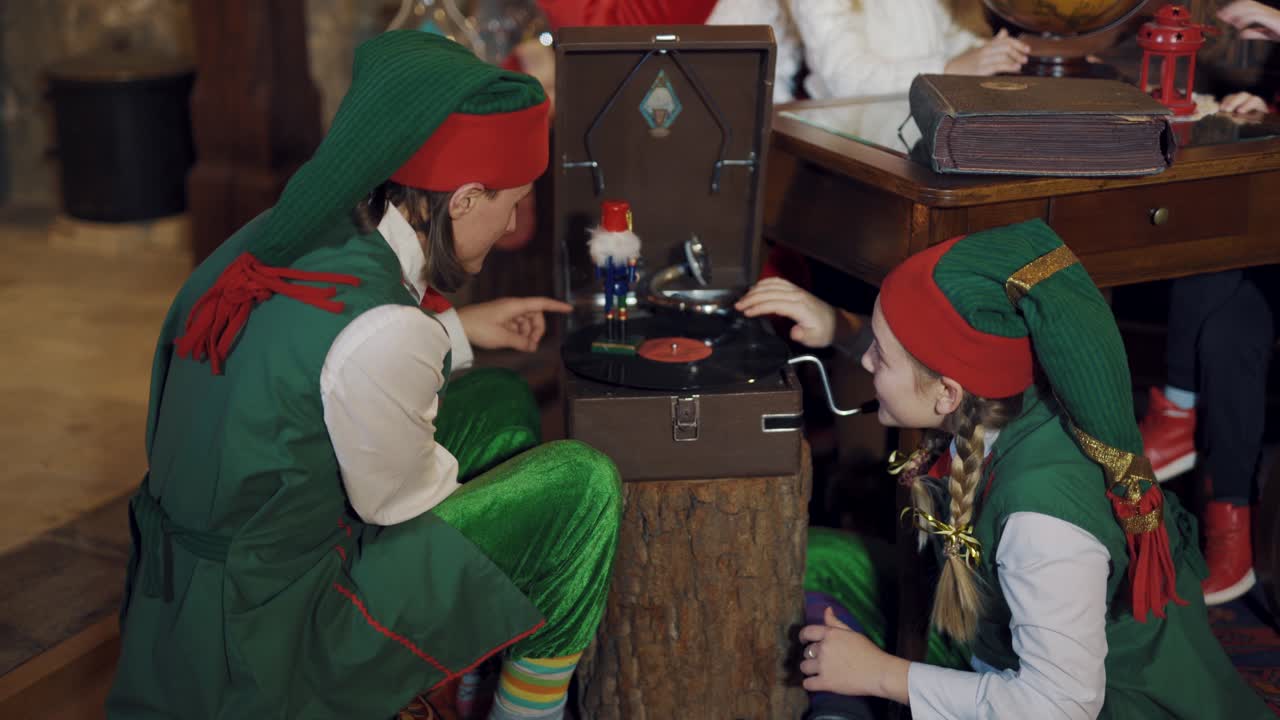 Funny elves in fairy costumes play with a toy indoors. Two elves male and female spinning a disk on the old recorder with toy soldier in Santa's residence.