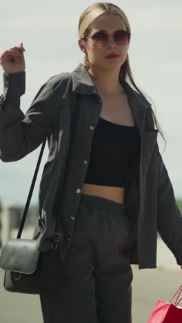 una dama con una camiseta negra bajo una chaqueta gris baila, chasqueando los dedos mientras sostiene bolsas de compras rojas, menta y verdes, con una colina a un lado y una vista borrosa de la gente caminando detrás de ella