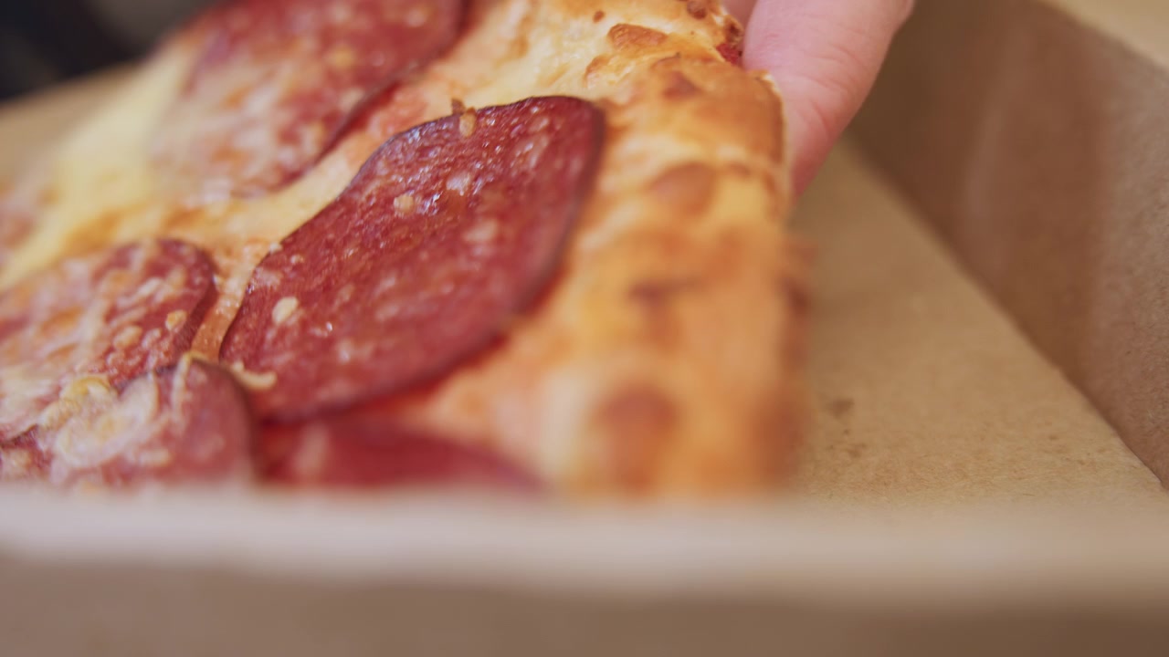 Kawałek pepperoni wyjmowany z pudełka, dłoń zabiera ostatni kawałek podczas swobodnej kolacji z dostawą, opuszki palców dotykają skórki, gdy okruszki spadają na karton, zbliżenie z płytką głębią ostrości uchwyca ruch i olej