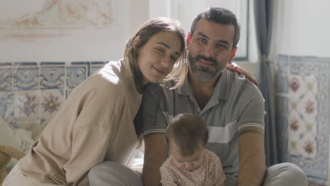 Portrait-Shot-Of-Happy-Family-Sitting-On-Bed-With-Their-Cute-Baby-Daughter-And-Looking-At-Camera