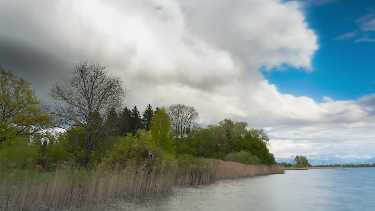 Dark clouds drift over a lake. Nature is beautiful, and the landscape blooming, for it is spring. A rainbow is visible, and everything is colorful. In the background, you can see the mountains.