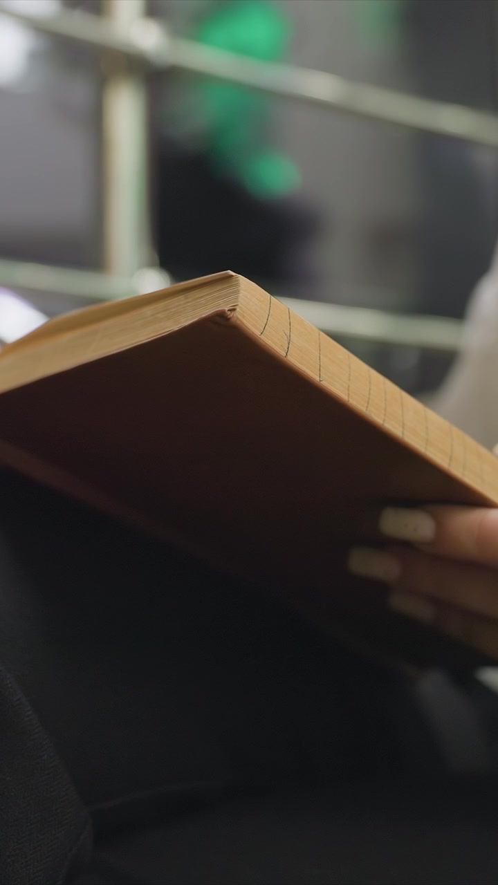 Primo piano di una mano che tiene un libro aperto con pagine visibili. Lo sfondo è sfocato e mostra forme indistinte. L'illuminazione soffusa evidenzia i dettagli di una tavola di legno
