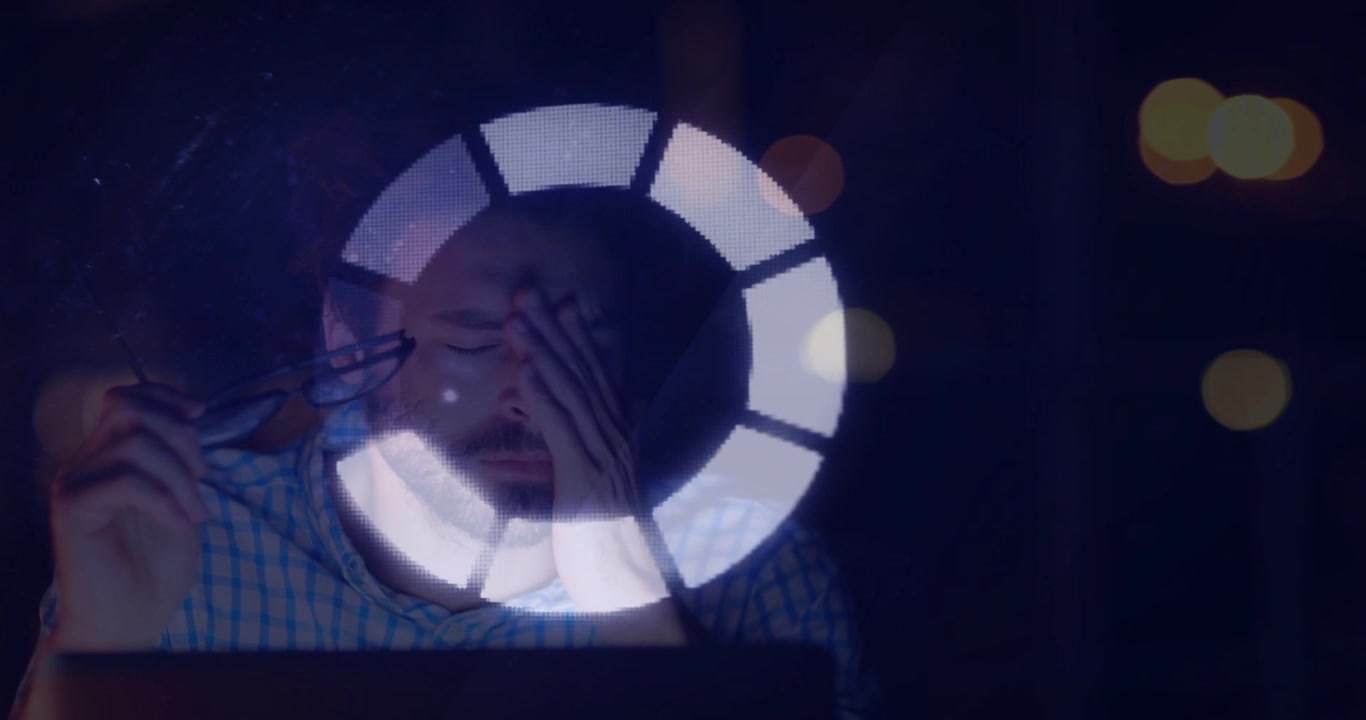 Man at laptop rubbing face, reacting to rotating ring reflection during late-night tech work