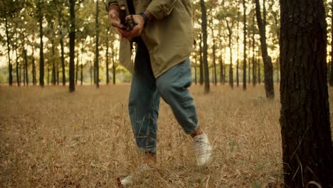 A-man-in-a-light-green-shirt-walks-through-a-forest-with-withered-grass-and-collects-firewood-to-start-a-fire.-Finding-firewood-for-a-fire-during-a-camping-stop