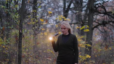 A-Woman-With-A-Flashlight-In-Hand-Walks-Through-The-Woods-At-Dusk