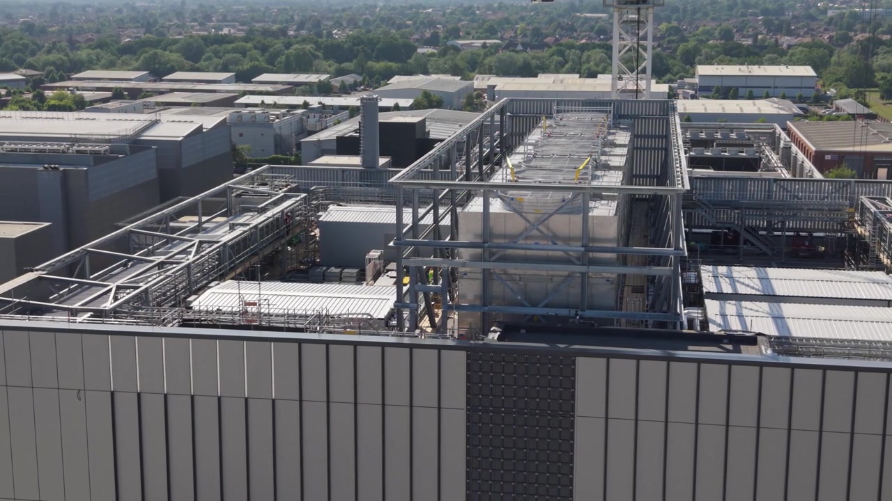 Vista aerea di un moderno edificio di data center con la sua estesa infrastruttura sul tetto in una giornata luminosa. Sono visibili complessi sistemi di raffreddamento, unità di ventilazione e apparecchiature di distribuzione dell'energia.