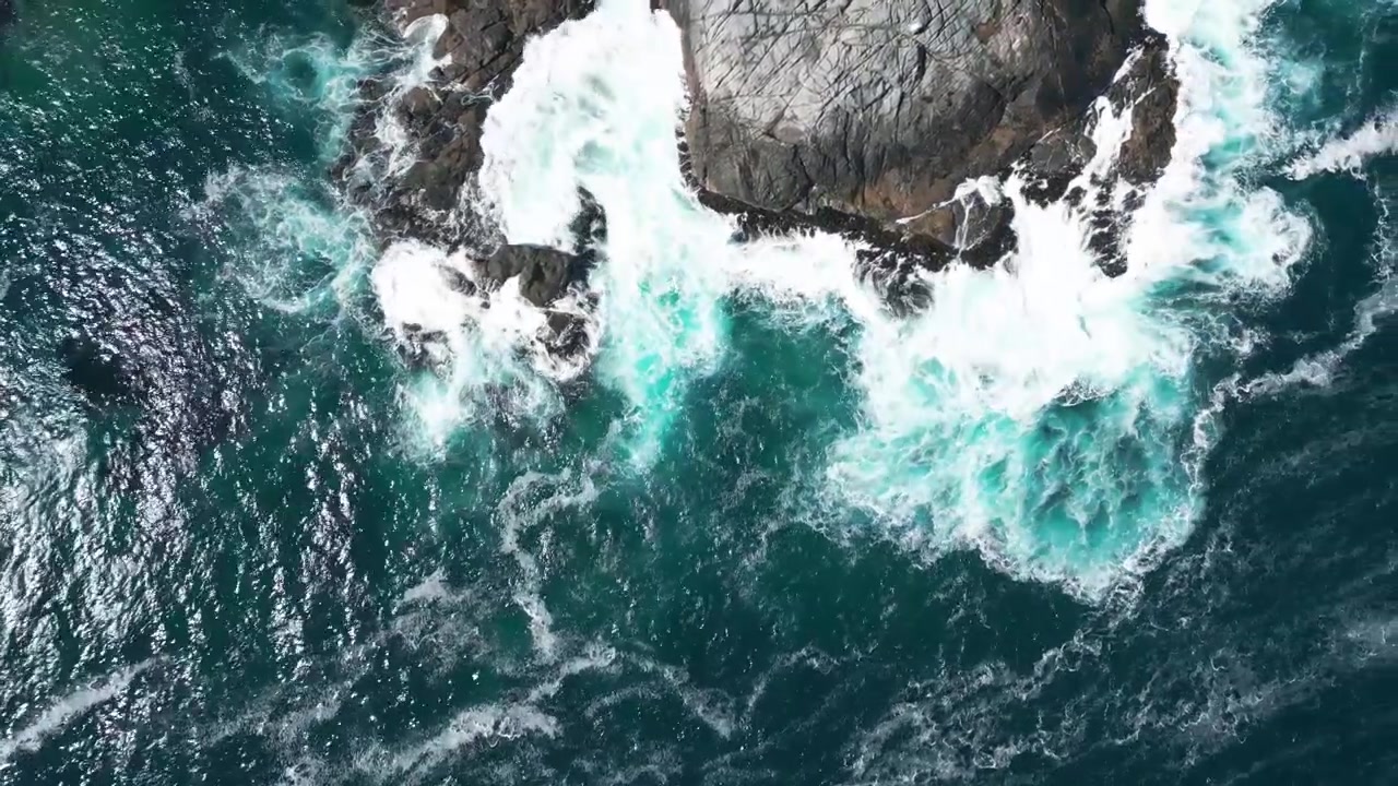 Top down view of turquoise foaming waves crashing against dark jagged rocks with white sea spray and deep blue swirling ocean below