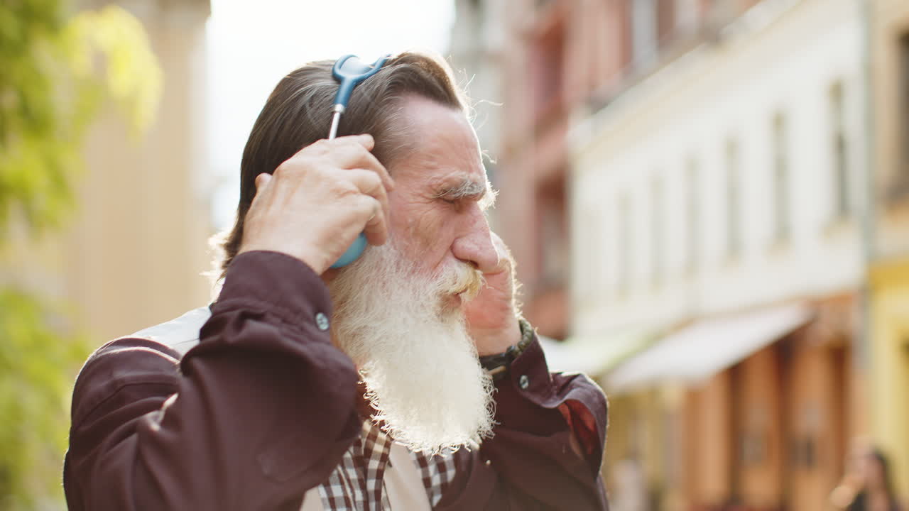 Happy senior old man in wireless headphones choosing listening music dancing outdoors city street