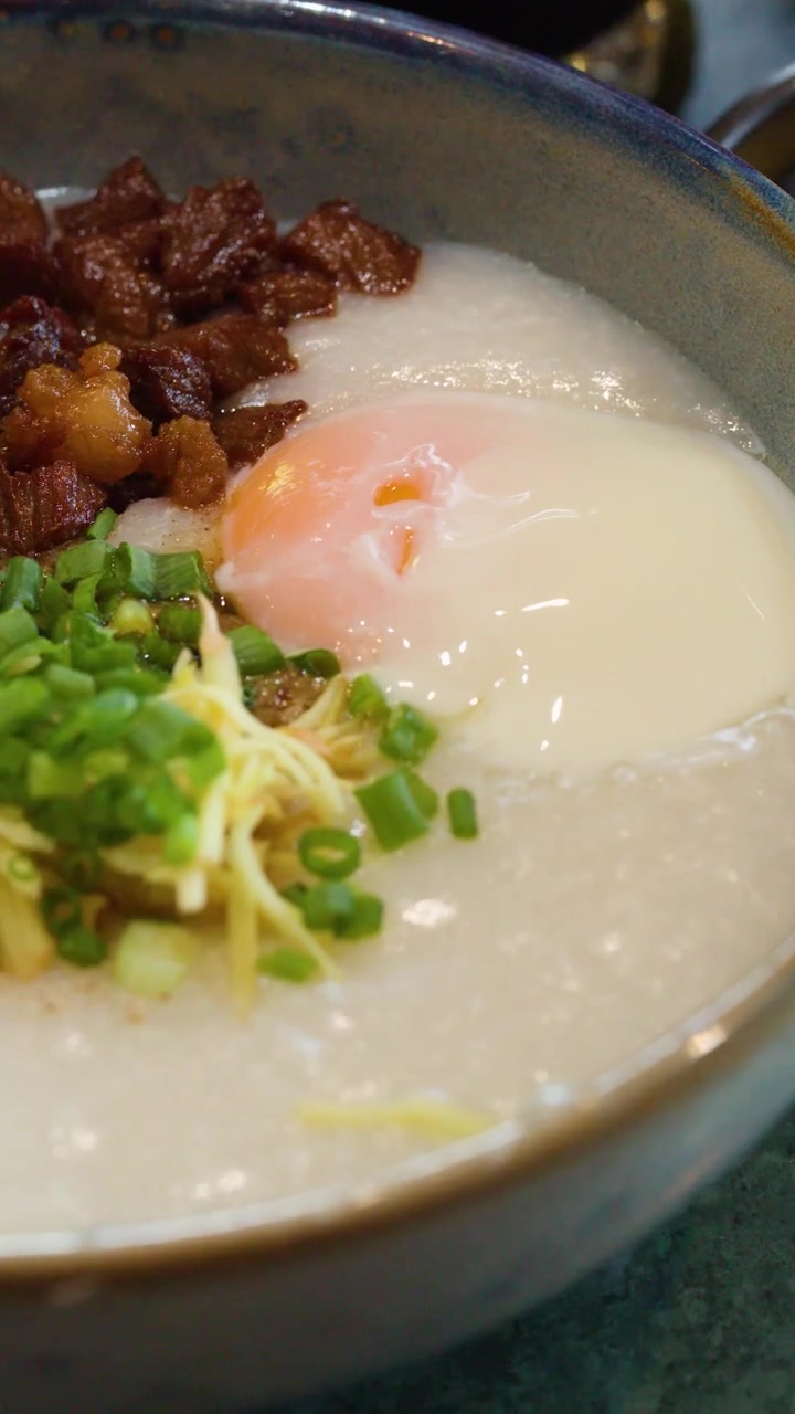 Kamera close-up bergerak melintasi mangkuk bubur nasi dengan topping telur, daging babi, dan daun bawang.