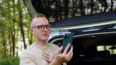 caucasian male in glasses looks at smartphone. man sitting in his car trunk talks on the phone and check his hair on the gadget screen.