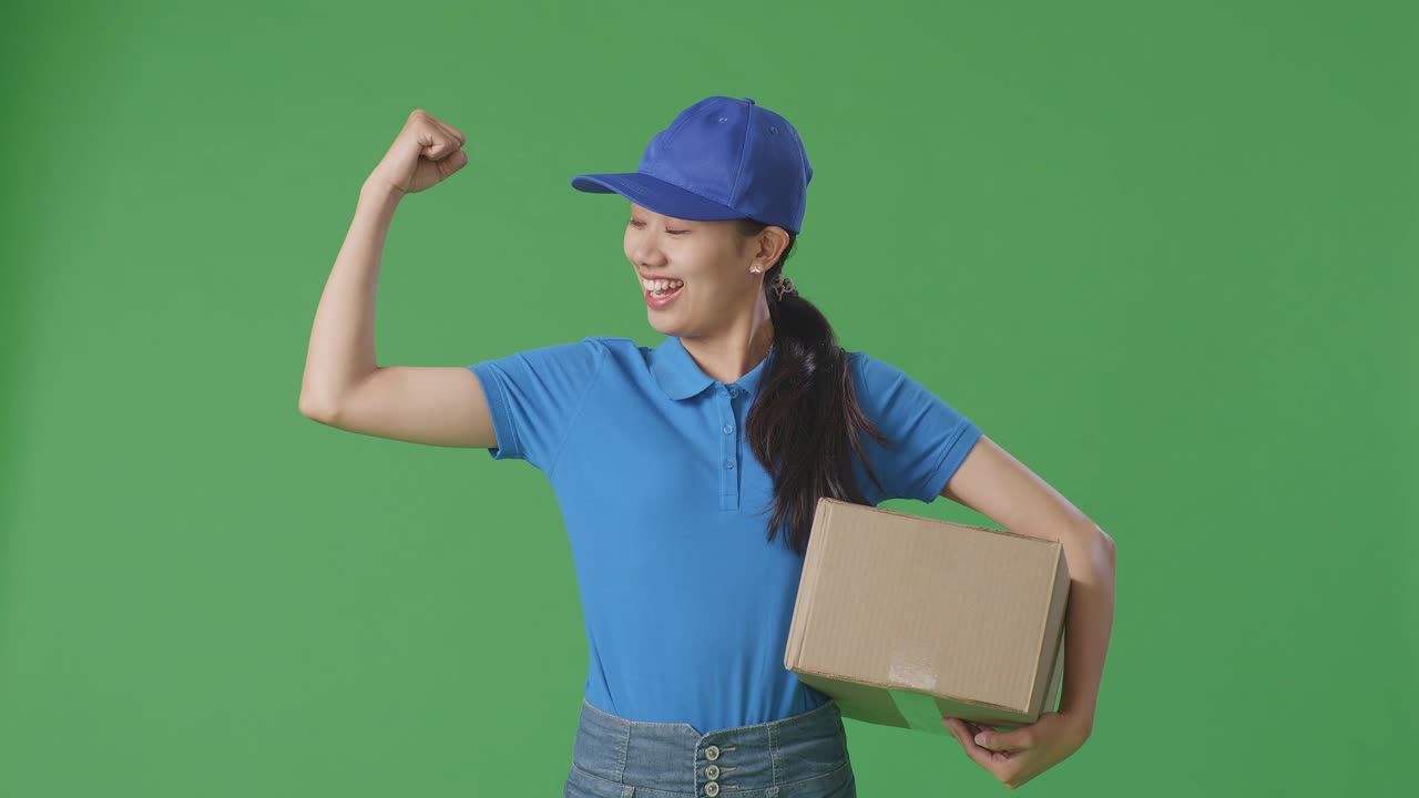 Asian Female Courier In Blue Uniform Smile And Flex Muscle While Delivering A Carton On Green Screen Background In The Studio