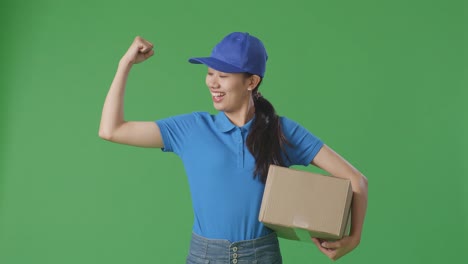 asian female courier in blue uniform smile and flex muscle while delivering a carton on green screen background in the studio