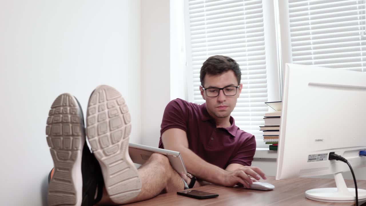 안경을 쓴 젊은 노동자가 다리에 키보드를  ⁇ 고 타이핑을 하고 다리가 테이블 위에 있는 사무실의 컴퓨터 화면을 보고 있다. 현대적인 사무실에서 바쁜 피곤한 노동자. 4k