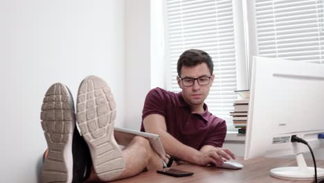 Joven-Trabajador-Con-Gafas-Sosteniendo-El-Teclado-En-Las-Piernas-Escribiendo-Y-Mirando-La-Pantalla-De-La-Computadora-En-La-Oficina-Con-Las-Piernas-Sobre-La-Mesa.-Trabajador-Cansado-Y-Ocupado-En-La-Oficina-Moderna.-4k