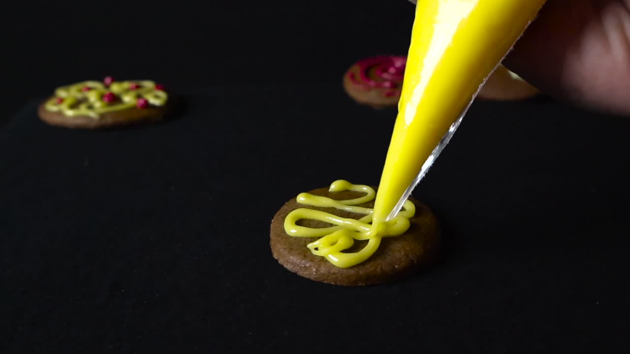 Caucasian white hand decorating gingerbread cookie with a sugar piping nozzle tip that has yellow vibrant colored sugar coating in it. The cookies are placed on a dark black studio background.