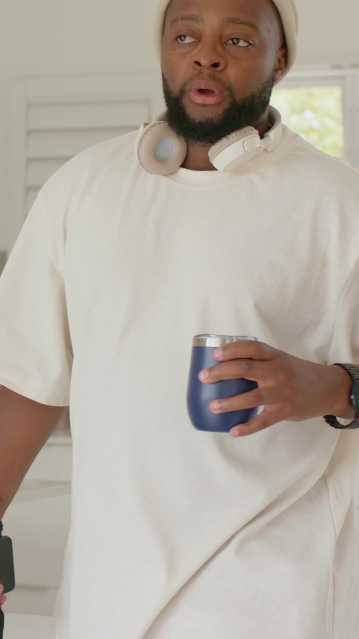 Video verticale: Uomo con berretto che allunga la mano per prendere una tazza blu, tenendo il telefono e spostando la borsa, mentre esce dalla cucina.