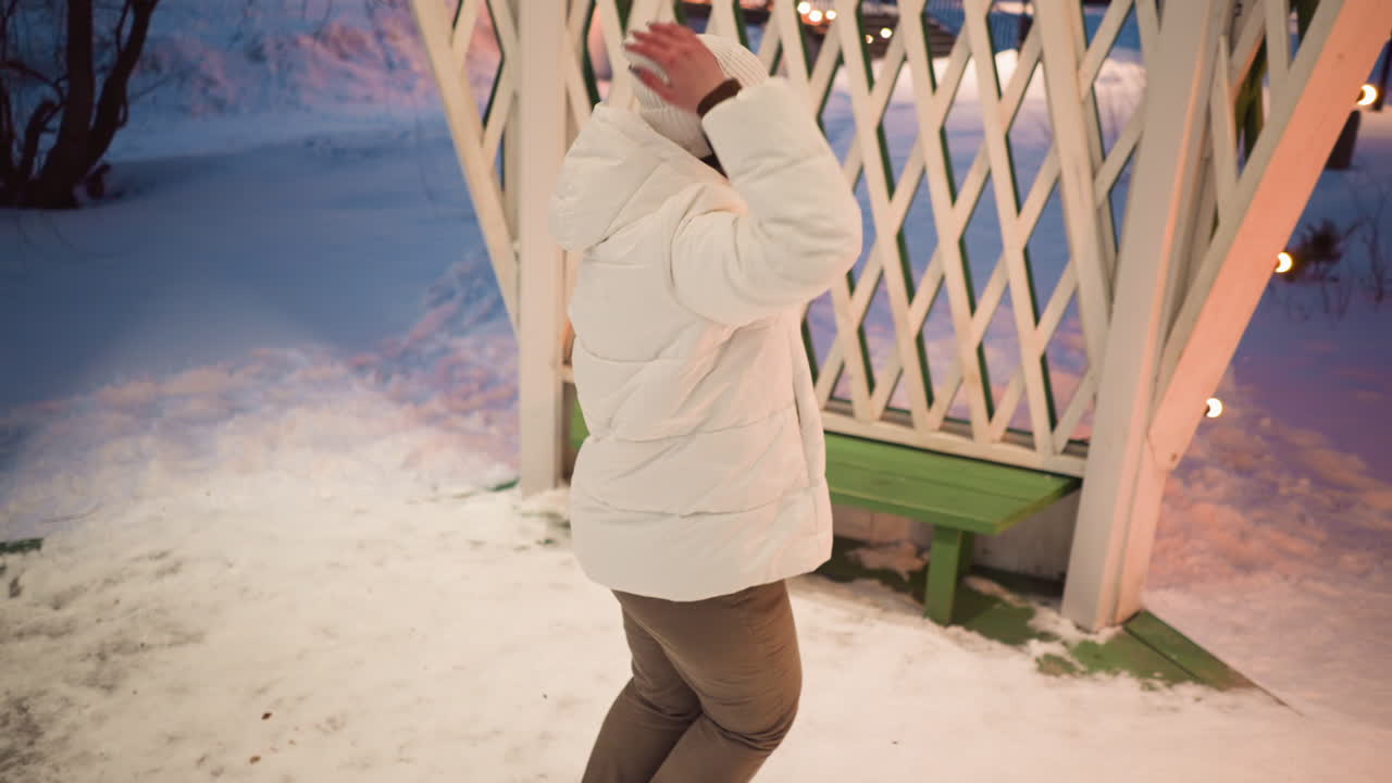 Back view of creative wearing headphones dancing to rhythm inside snow covered gazebo lit by bulbs under evening sky wearing puffer coat and beanie as gentle breeze tousles hair around bare trees