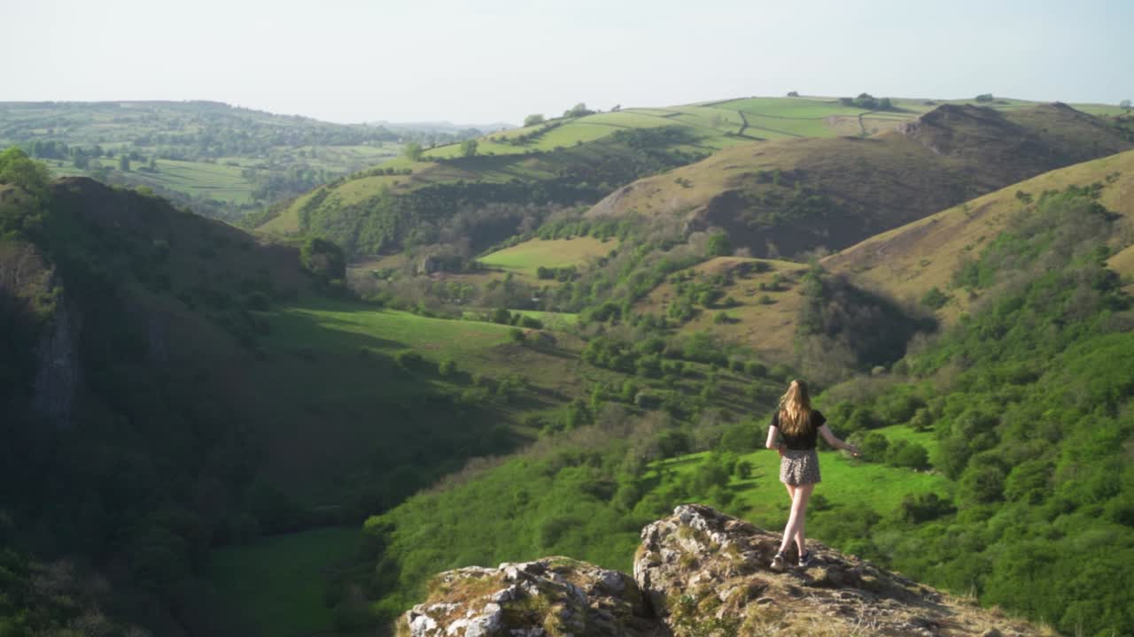 de mano, ancha, cámara lenta de una joven rubia de pie en el borde del acantilado sobre la cueva de thor, ashbourne, distrito de los picos, inglaterra al atardecer