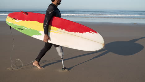 side view of a male surfer with artificial leg walking along beach and holding surfboard under arm