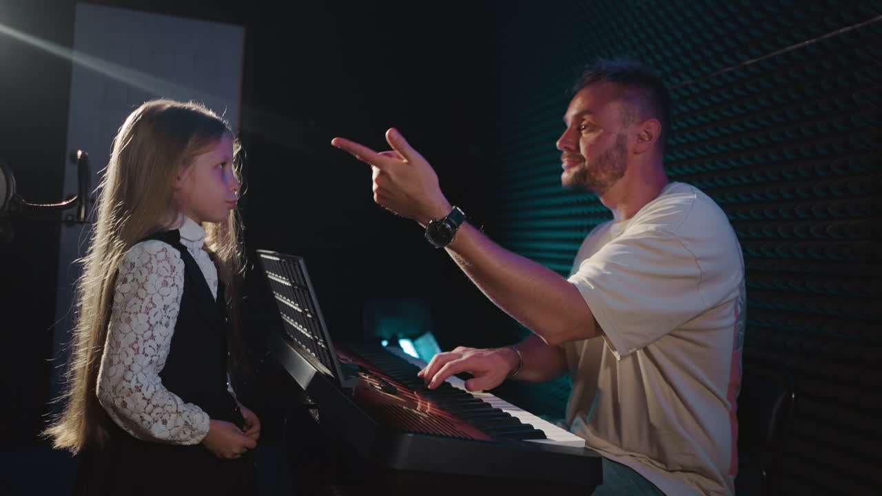 학교 소녀는 오디오 스튜디오에서 코치와 협력합니다. 신사는 보컬 연습에 참여하는 어린 소녀와 함께 전자 피아노를 연주합니다. 보컬 훈련 세션