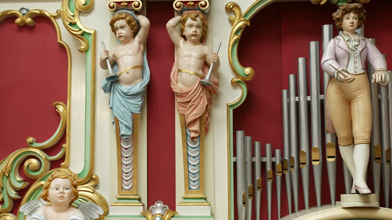 Statues on a decorative organ are standing between organ pipes, holding instruments in a symmetrical layout. Baroque-style figures and ornate carvings frame the vintage musical installation