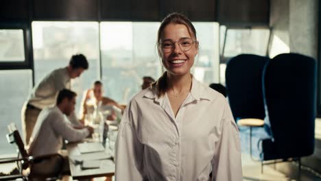 portrait of a happy girl in glasses and a white shirt in the office, who smiles and looks at the camera. posing against the backdrop of the working process of office workers