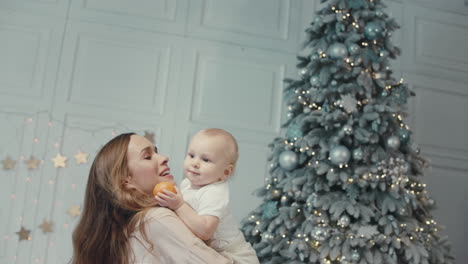 cheerful mother kissing cute child near christmas tree in modern apartment.