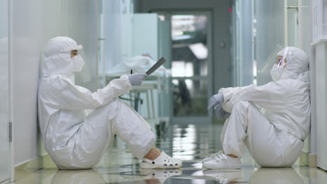 Two-Female-Doctors-in-Protective-Wear-Resting-on-Floor-in-Corridor
