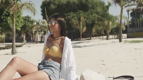 mujer hispana feliz con gafas de sol sentada en la playa al sol, espacio de copia, cámara lenta