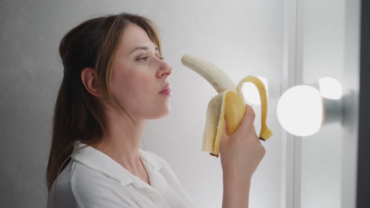 young woman standing front mirror eating banana observing reflection thoughtful mood adjusting hair bright vanity lights gentle illumination casual atmosphere beauty wellness lifestyle scene