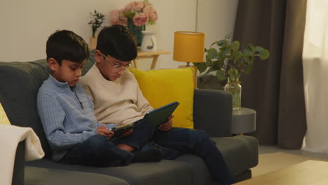 two young boys sitting on sofa at home playing games or streaming onto digital tablets 10