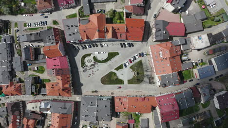 Top-down-view-of-market-square-in-old-European-town-surrounded-by-tenement-houses,-people-gathered-at-the-fountain,-parking-places-occupied