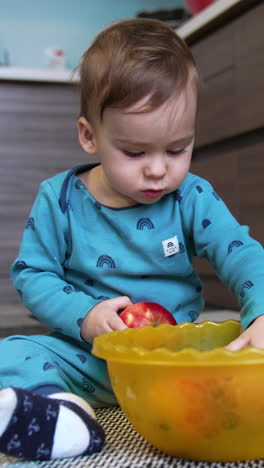 cute toddler sitting on the kitchen floor with a bowl of fruit in front of him. lovely child takes and apple and bites it looking at camera.