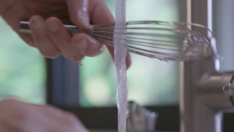 a caucasian male is cleaning and rinsing a french whisk in a home kitchen