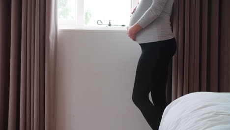 Smiling-Pregnant-Woman-Standing-By-Window-Holding-Belly