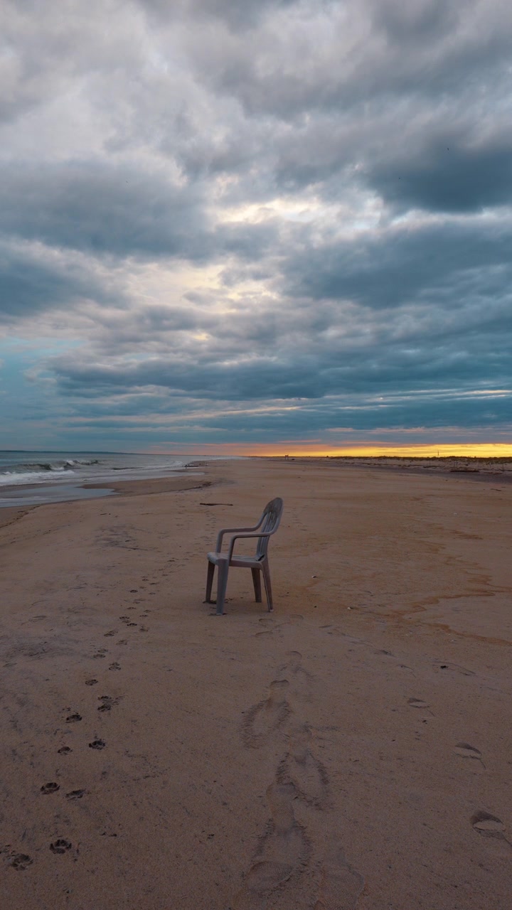 Chair on the beach near the water at sunset with clouds in the sky. A plastic chair sits alone on the sand by the water as the sun sets.
