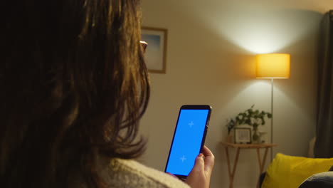 rear view of young woman spending evening at home sitting on sofa looking at blue screen mobile phone