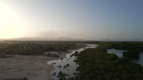 Drone-follows-an-extended-body-of-water,-mangrove-forest,-long-sandbanks-on-the-left,-river-bends-to-the-horizon-showing-the-sun-setting-and-mountains,-Porlamar,-Margarita-Island,-Venezuela
