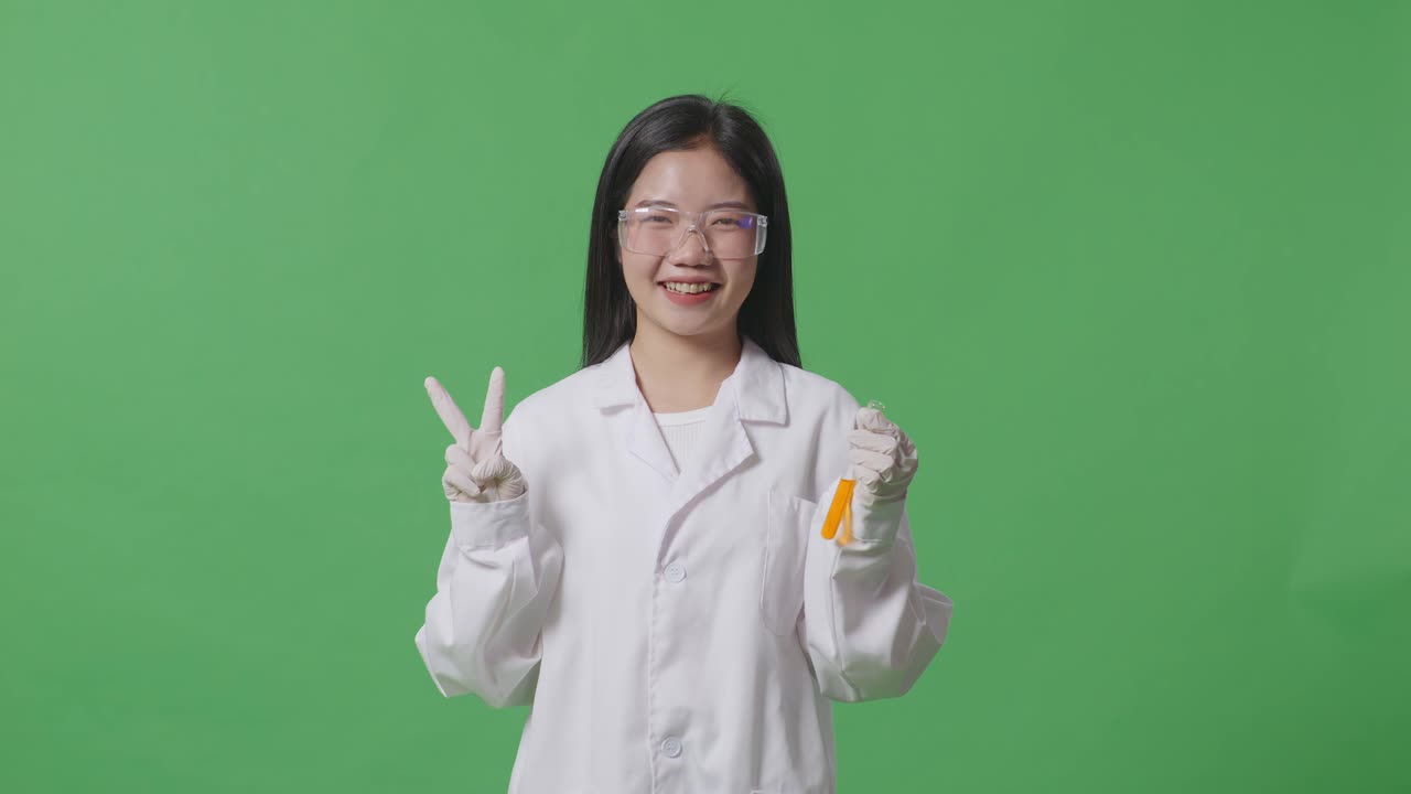 실험실에서 녹색 스크린 배경에 서 있는 동안 미소 짓고 평화의 몸짓을 보여주는 시험관에서 오렌지 액체와 함께 아시아 여성 과학자
