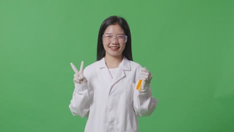 실험실에서 녹색 스크린 배경에 서 있는 동안 미소 짓고 평화의 몸짓을 보여주는 시험관에서 오렌지 액체와 함께 아시아 여성 과학자