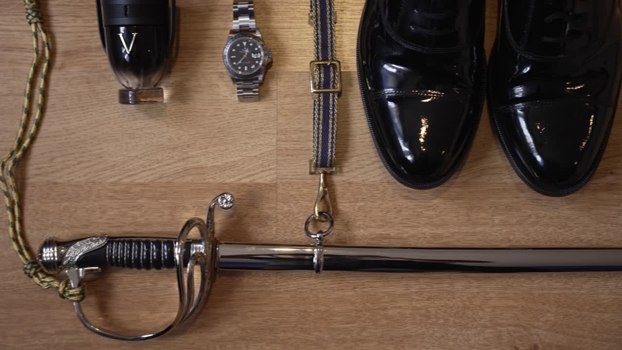 Formal accessories neatly arranged: polished shoes, watch, belt, sword, and cologne