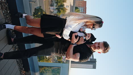 young parents of a newborn boy stand together on top of the building. mother waves her baby and husband smiles to her. transport runs at backdrop. vertical view.