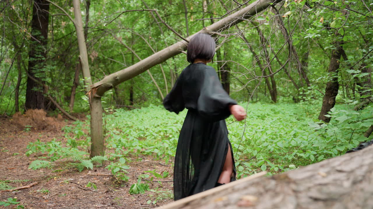 Woman wearing black flowing dress turns around gracefully in green forest near fallen tree, movement of sheer fabric blending with soft natural light, creating dreamy cinematic scene
