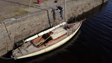 man climbs down ladder from rock wall onto damaged sailboat docked at stone pier