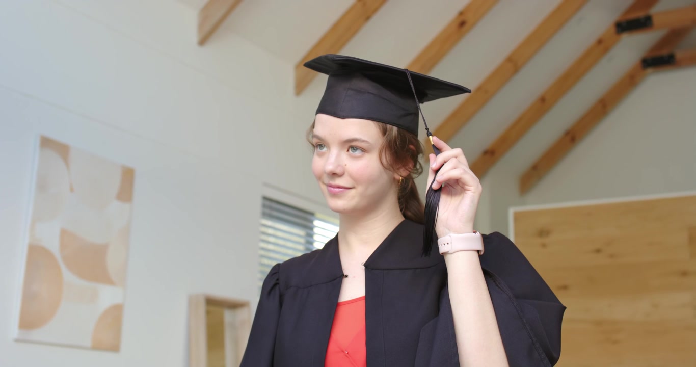 Donna che si sistema il tocco, tenendo la nappa dopo averla posizionata, preparandosi per la laurea in una stanza con travi in legno