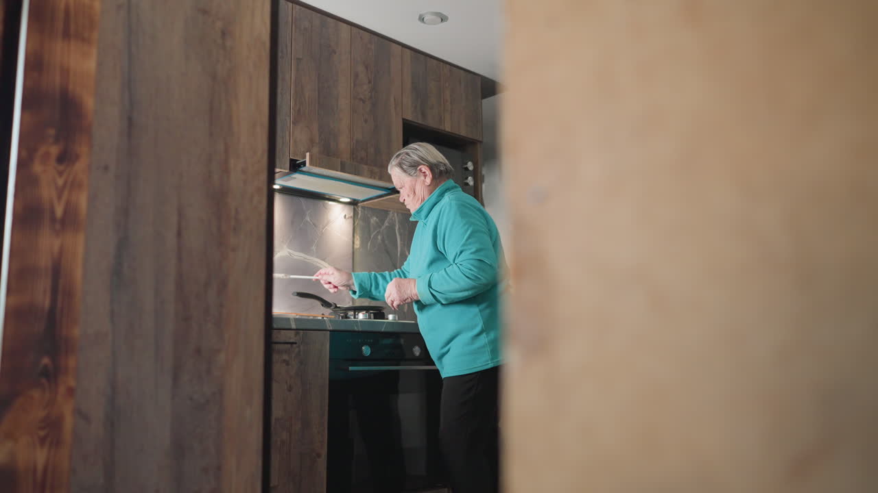 Older woman standing in modern kitchen, preparing meal, focused on cooking process, holding food ingredient while looking at stove, with kitchen tools, stovetop, and fridge in background