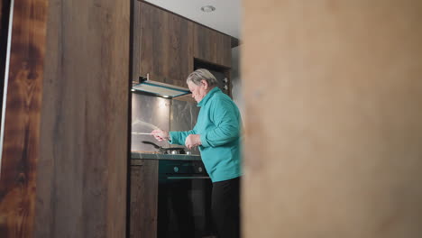 older woman standing in modern kitchen, preparing meal, focused on cooking process, holding food ingredient while looking at stove, with kitchen tools, stovetop, and fridge in background