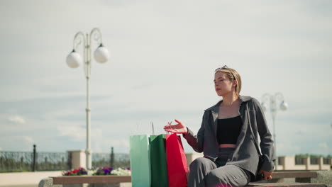 lady places her shopping bags on a bench, sits down, and relaxes her left hand while sunlight reflects on her face, in the blurred background, a cyclist rides by as a street lamp is visible